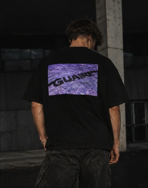 From behind: the tee's back print, GUAIRE across a purple, textured grass photo.