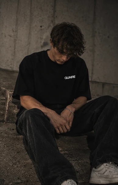 Sitting on a concrete step, head down, in the black Guaire tee and wide black trousers.